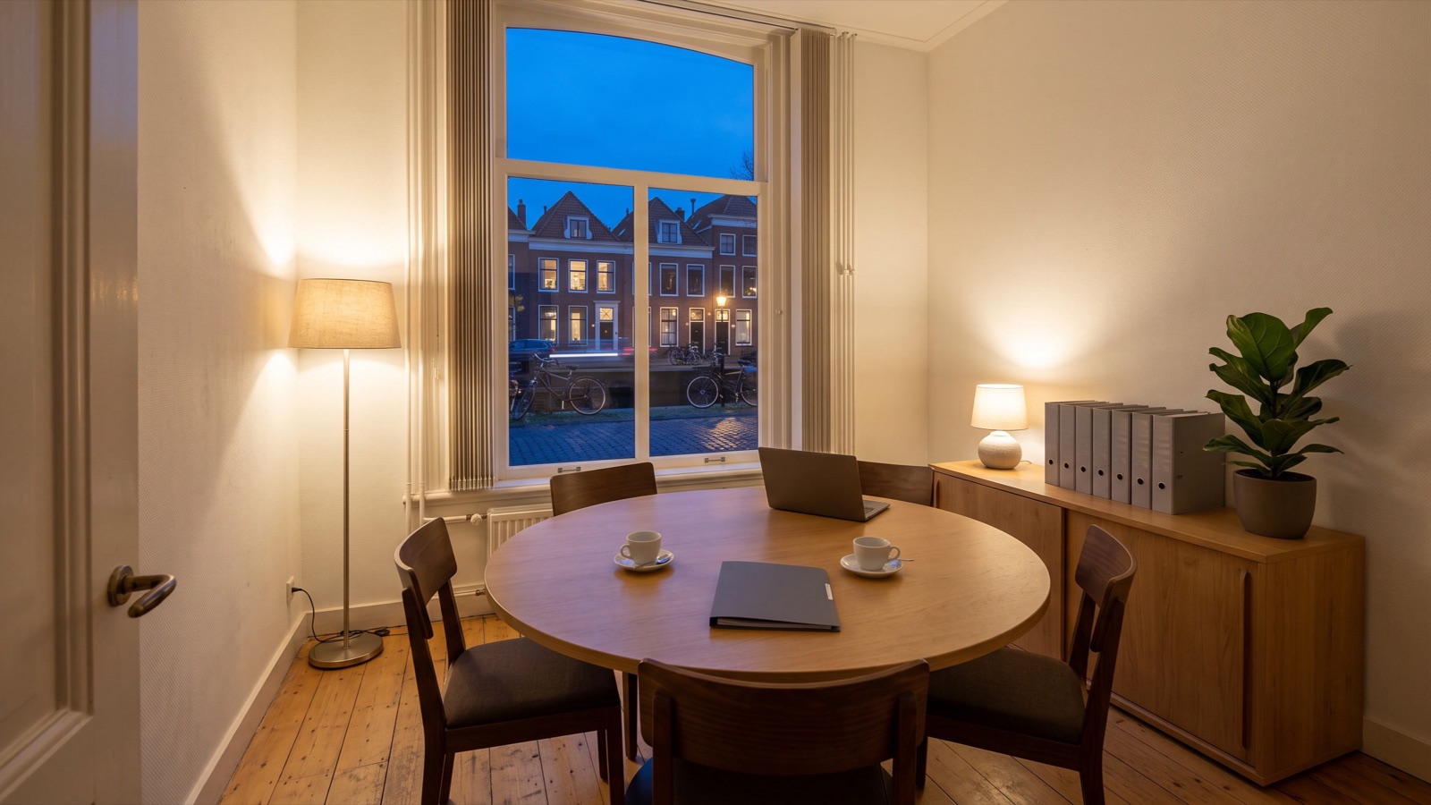 De lege spreekkamer van een adviespraktijk aan het eind van de dag, met lamplicht en een gracht buiten