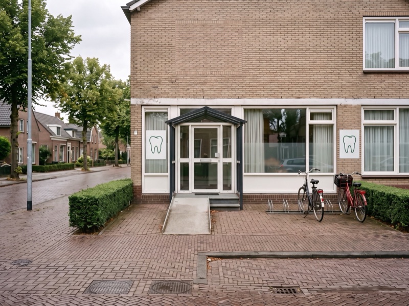 De voorkant van de praktijk aan een rustige straat, met een glazen entree, twee witte borden met een tandsymbool en een hellingbaan naar de deur